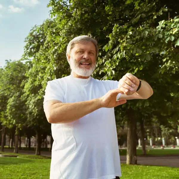 Smiling older man with a white beard in a white T-shirt, standing in a sunny park and looking at his wrist